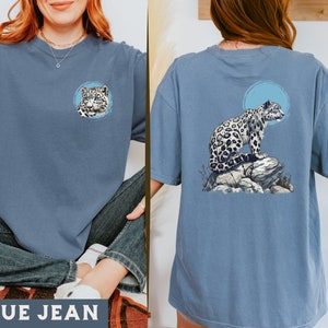 May include: A blue jean t-shirt featuring a snow leopard graphic on the front and back. The front graphic is a close-up of the snow leopard's face, while the back graphic shows the snow leopard sitting on a rock.
