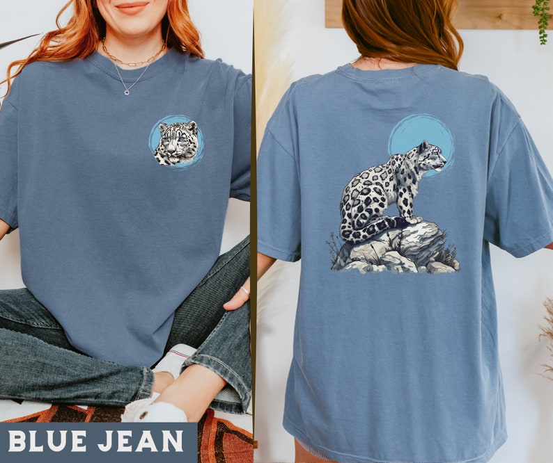 May include: A blue jean t-shirt featuring a snow leopard graphic on the front and back. The front graphic is a close-up of the snow leopard's face, while the back graphic shows the snow leopard sitting on a rock.