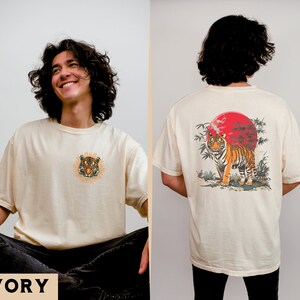 Op de afbeelding: Een cr&egrave;mekleurig T-shirt met een tijgerprint aan de voorkant en een tijgerprint met een rode zon en bamboe aan de achterkant. De tekst "IVORY" staat onderaan de afbeelding.