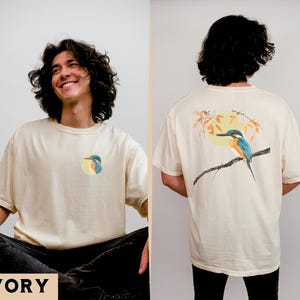 Puede incluir: Una camiseta de color crema con un gr&aacute;fico de un p&aacute;jaro azul y verde en la parte delantera y una versi&oacute;n m&aacute;s grande del mismo gr&aacute;fico de p&aacute;jaro en la parte posterior. El texto "IVORY" est&aacute; impreso en la parte inferior de la imagen.