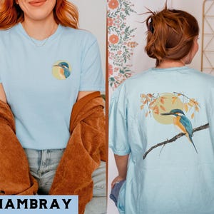 Puede incluir: Dos camisetas de color azul claro con un dise&ntilde;o de p&aacute;jaro. Una camiseta presenta un mart&iacute;n pescador en una rama con hojas y un sol. La otra camiseta presenta un mart&iacute;n pescador sobre un fondo azul.