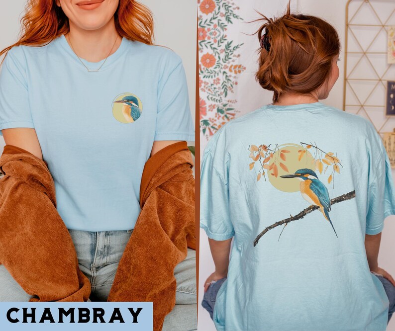 Puede incluir: Dos camisetas de color azul claro con un dise&ntilde;o de p&aacute;jaro. Una camiseta presenta un mart&iacute;n pescador en una rama con hojas y un sol. La otra camiseta presenta un mart&iacute;n pescador sobre un fondo azul.