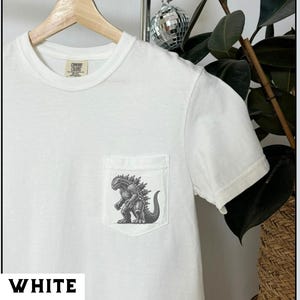 May include: White t-shirt with a pocket featuring a gray illustration of a Godzilla-like creature. The shirt is made of a soft material and has a crew neck. The word "WHITE" is printed at the bottom of the image.