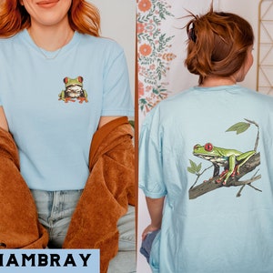 May include: A light blue t-shirt with a green tree frog on the front and a larger green tree frog on the back. The frog on the back is sitting on a branch with leaves.