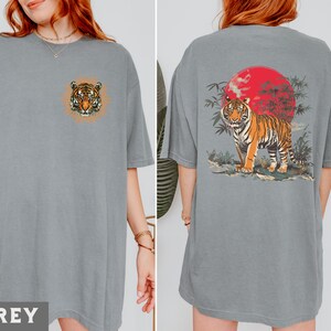 Op de afbeelding: Twee grijze T-shirts met een tijgerprint. E&eacute;n shirt heeft een tijgerkop met een bloemenontwerp, het andere shirt heeft een tijger in een jungle sc&egrave;ne met een rode zon.