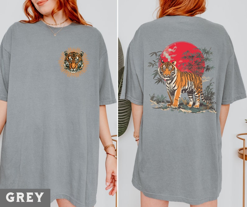 Op de afbeelding: Twee grijze T-shirts met een tijgerprint. E&eacute;n shirt heeft een tijgerkop met een bloemenontwerp, het andere shirt heeft een tijger in een jungle sc&egrave;ne met een rode zon.