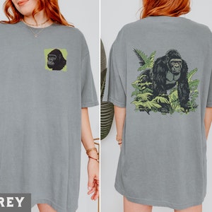 May include: A grey t-shirt with a gorilla graphic on the front. The gorilla is surrounded by green leaves. The shirt is a relaxed fit and has a crew neck.