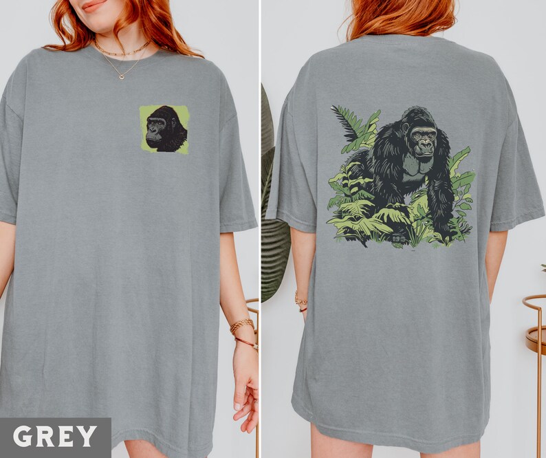 May include: A grey t-shirt with a gorilla graphic on the front. The gorilla is surrounded by green leaves. The shirt is a relaxed fit and has a crew neck.