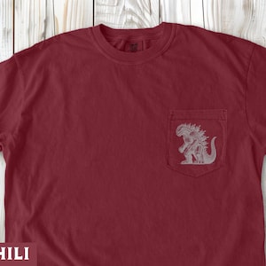 May include: A chili-colored t-shirt with a pocket featuring a gray Godzilla graphic. The shirt has a crew neck and short sleeves. The word "CHILI" is at the bottom of the image.