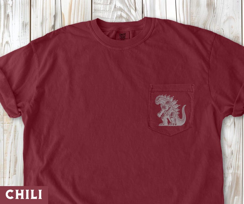 May include: A chili-colored t-shirt with a pocket featuring a gray Godzilla graphic. The shirt has a crew neck and short sleeves. The word "CHILI" is at the bottom of the image.
