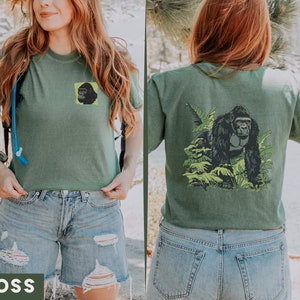 May include: A green t-shirt with a gorilla graphic on the back and a smaller gorilla graphic on the front. The shirt is being worn by a person with long brown hair. The person is wearing blue denim shorts.