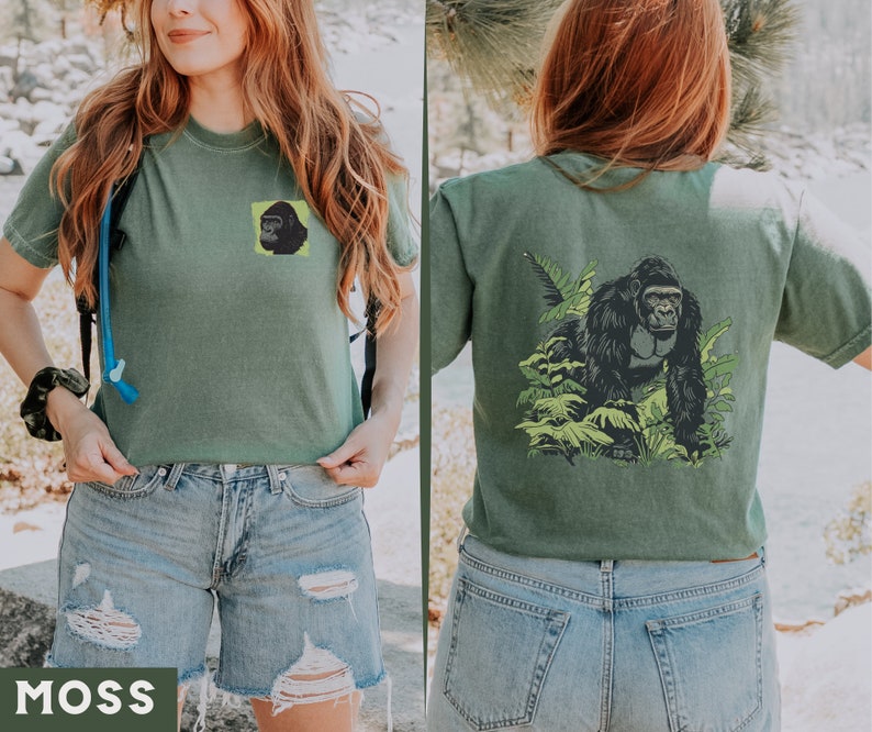 May include: A green t-shirt with a gorilla graphic on the back and a smaller gorilla graphic on the front. The shirt is being worn by a person with long brown hair. The person is wearing blue denim shorts.