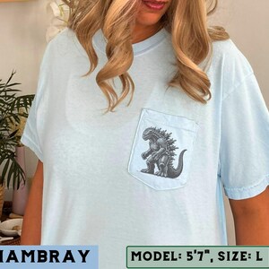 May include: Light blue chambray t-shirt with a pocket featuring a Godzilla graphic. The graphic is a detailed black and white illustration of the iconic monster. The shirt has short sleeves and a relaxed fit, perfect for casual wear.
