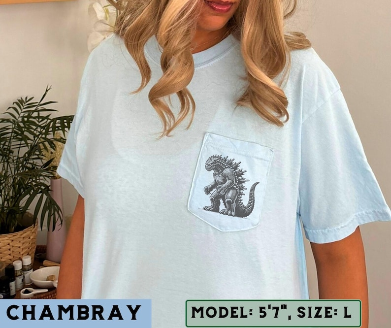 May include: Light blue chambray t-shirt with a pocket featuring a Godzilla graphic. The graphic is a detailed black and white illustration of the iconic monster. The shirt has short sleeves and a relaxed fit, perfect for casual wear.