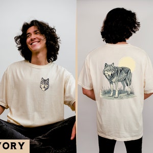 May include: A cream colored t-shirt with a wolf graphic on the front and a wolf graphic with a full moon on the back. The text "IVORY" is printed on the bottom of the image.