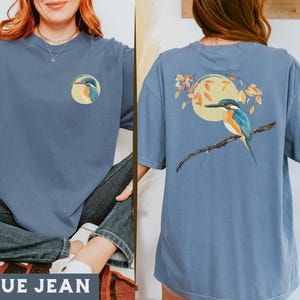 Puede incluir: Una camiseta azul jean con un gr&aacute;fico de p&aacute;jaro en la parte delantera y un gr&aacute;fico de p&aacute;jaro en la parte posterior. El p&aacute;jaro es un mart&iacute;n pescador con plumas azules, verdes y naranjas. La camiseta tiene un corte holgado y cuello redondo.