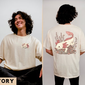 May include: A cream colored t-shirt with a small axolotl graphic on the front and a larger axolotl graphic on the back. The axolotl is pink and white with black eyes and is surrounded by pink flowers. The text "IVORY" is printed in black at the bottom of the image.