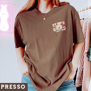 May include: A brown t-shirt with a graphic of an axolotl on the front. The axolotl is a pink and white amphibian with a cute expression.
