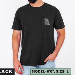 May include: Black t-shirt with a pocket featuring a detailed graphic of a monster. The shirt is a solid black color. The text "BLACK" is at the bottom left of the image. The text "MODEL: 6'0", SIZE: L" is at the bottom right.
