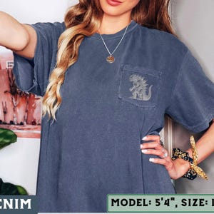 May include: Denim blue t-shirt with a pocket featuring a gray graphic of a creature. The shirt has short sleeves and a relaxed fit. The text "DENIM" is at the bottom left. The model is 5'4" and wearing a size L.