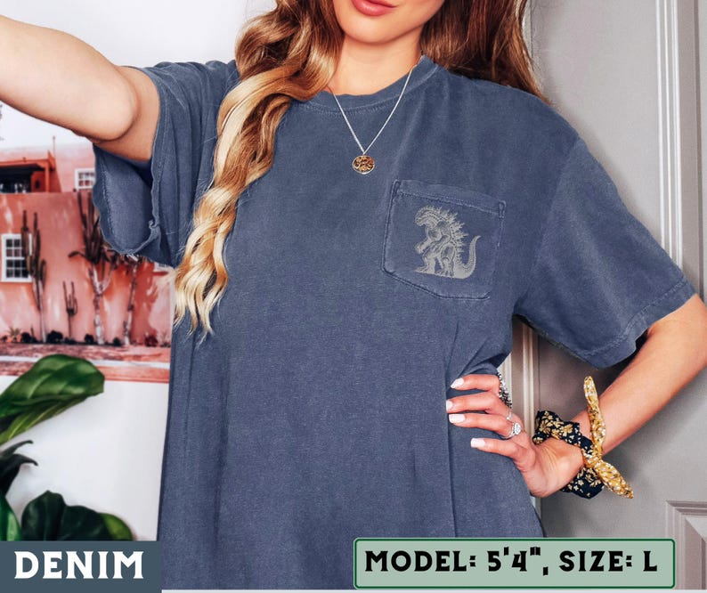 May include: Denim blue t-shirt with a pocket featuring a gray graphic of a creature. The shirt has short sleeves and a relaxed fit. The text "DENIM" is at the bottom left. The model is 5'4" and wearing a size L.