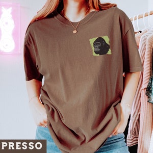 May include: A brown t-shirt with a green and black gorilla graphic on the front. The shirt is being worn by a person with long brown hair.