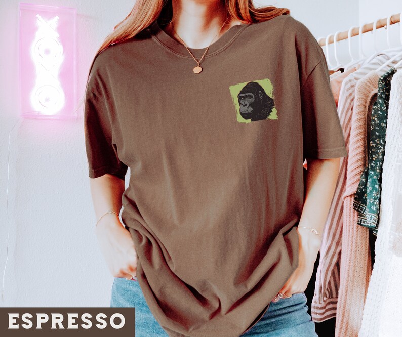 May include: A brown t-shirt with a green and black gorilla graphic on the front. The shirt is being worn by a person with long brown hair.
