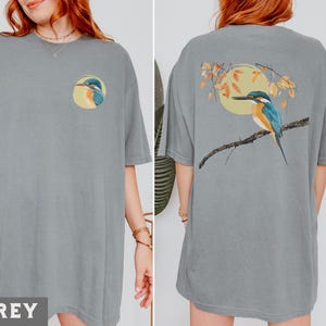 Puede incluir: Una camiseta gris de gran tama&ntilde;o con un gr&aacute;fico de un p&aacute;jaro en la parte delantera y un gr&aacute;fico de un p&aacute;jaro en la parte trasera. El gr&aacute;fico del p&aacute;jaro en la parte delantera es un primer plano de la cabeza del p&aacute;jaro. El gr&aacute;fico del p&aacute;jaro en la parte trasera muestra al p&aacute;jaro posado en una rama con hojas.