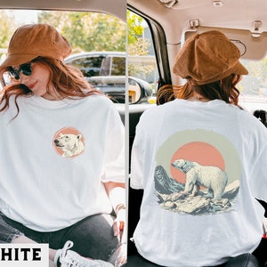 May include: White t-shirt with a graphic of a polar bear in a mountain landscape with a red sun. The graphic is on the back of the shirt. The shirt is being worn by a person.