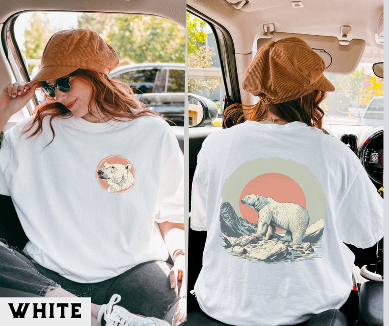 May include: White t-shirt with a graphic of a polar bear in a mountain landscape with a red sun. The graphic is on the back of the shirt. The shirt is being worn by a person.