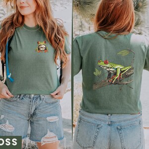 May include: A green t-shirt with a green frog graphic on the front and a larger green frog graphic on the back. The frog has red eyes and is sitting on a branch. The shirt is a cropped style and is being worn by a person with blue jeans.
