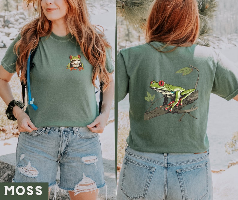 May include: A green t-shirt with a green frog graphic on the front and a larger green frog graphic on the back. The frog has red eyes and is sitting on a branch. The shirt is a cropped style and is being worn by a person with blue jeans.