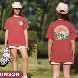 May include: A red t-shirt with a white polar bear graphic on the front and a white polar bear graphic on the back. The shirt has a crew neck and short sleeves. The polar bear graphic on the back is surrounded by a circle with a red sun and blue mountains.