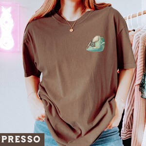 May include: A brown t-shirt with a green chameleon graphic on the front. The chameleon is facing left and has a light green body with dark green spots. The t-shirt is a relaxed fit and has a crew neck.