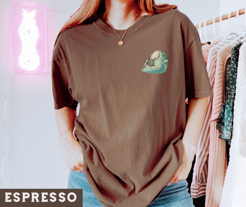 May include: A brown t-shirt with a green chameleon graphic on the front. The chameleon is facing left and has a light green body with dark green spots. The t-shirt is a relaxed fit and has a crew neck.