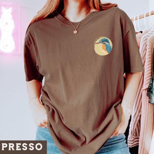 Puede incluir: Una camiseta marr&oacute;n de manga corta con un gr&aacute;fico de un mart&iacute;n pescador azul y verde en un c&iacute;rculo. La camiseta tiene cuello redondo y est&aacute; hecha de una tela suave y c&oacute;moda.