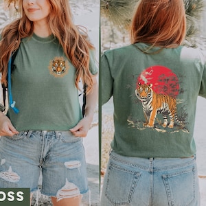 Op de afbeelding: Een groen T-shirt met een tijgerprint aan de voorkant en een tijgerprint met een rode zon en palmbomen aan de achterkant. Het shirt wordt gedragen door een persoon met lang bruin haar en een blauwe jeans.