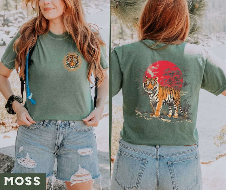 Op de afbeelding: Een groen T-shirt met een tijgerprint aan de voorkant en een tijgerprint met een rode zon en palmbomen aan de achterkant. Het shirt wordt gedragen door een persoon met lang bruin haar en een blauwe jeans.