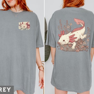 May include: A grey t-shirt with a pink and white axolotl illustration on the front. The axolotl is surrounded by pink flowers and leaves.