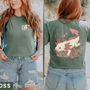 May include: A green t-shirt with a graphic of an axolotl on the back. The axolotl is pink and white with red gills. The shirt is cropped and has a distressed look. The model is wearing blue jeans.