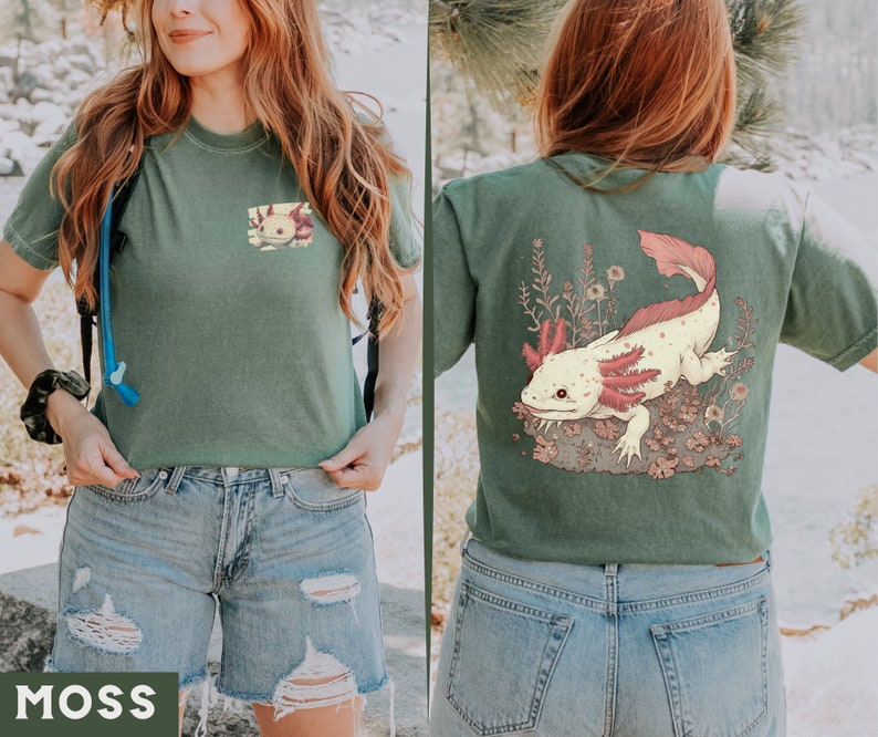 May include: A green t-shirt with a graphic of an axolotl on the back. The axolotl is pink and white with red gills. The shirt is cropped and has a distressed look. The model is wearing blue jeans.