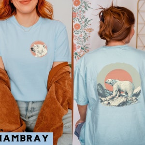 May include: Two light blue t-shirts with a polar bear graphic. One shirt features a polar bear head in a circle, the other features a polar bear walking in a landscape with a sunset.
