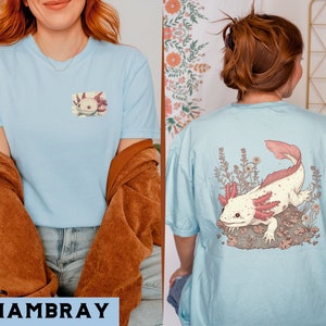 May include: Two light blue t-shirts with a pink axolotl illustration on the back. The axolotl is surrounded by green and white flowers. The t-shirts are made of a soft, comfortable fabric and are perfect for everyday wear.