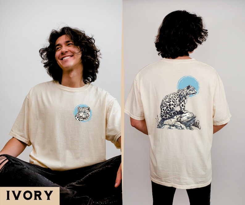 May include: A cream coloured t-shirt with a snow leopard graphic on the front and a larger snow leopard graphic on the back. The snow leopard is sitting on a rock with a blue circle behind it. The text "IVORY" is printed in black at the bottom of the image.