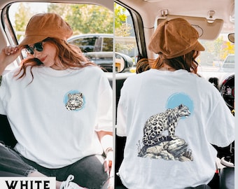 以下が含まれることがあります： 白地に雪豹のグラフィックがプリントされたTシャツ。前面の雪豹は青い背景の円の中に描かれており、背面の雪豹は青い円を背景に岩の上に立っている。