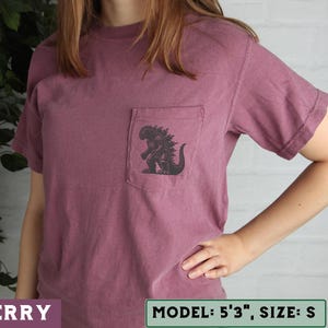 May include: Berry-colored short-sleeve t-shirt with a pocket featuring a black Godzilla graphic. The shirt is a size small. The model is 5'3".