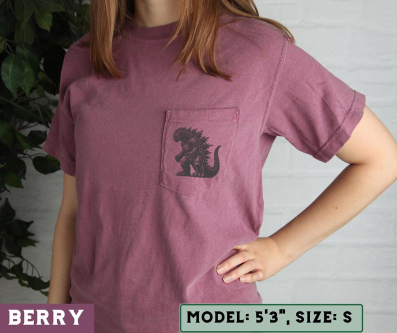 May include: Berry-colored short-sleeve t-shirt with a pocket featuring a black Godzilla graphic. The shirt is a size small. The model is 5'3".
