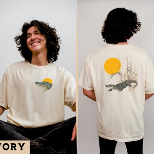 Op de afbeelding: Een crèmekleurig T-shirt met een afbeelding van een alligator in een moeras met een gele zon. De achterkant van het shirt heeft een vergelijkbare afbeelding met de alligator in het water met hoog gras en een gele zon. De tekst "IVORY" is in zwart gedrukt aan de onderkant van de afbeelding.