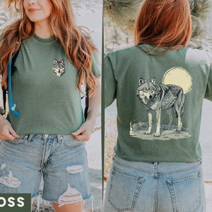 May include: A green t-shirt with a wolf graphic on the front and a wolf graphic with a full moon on the back. The model is wearing the shirt with blue denim shorts.