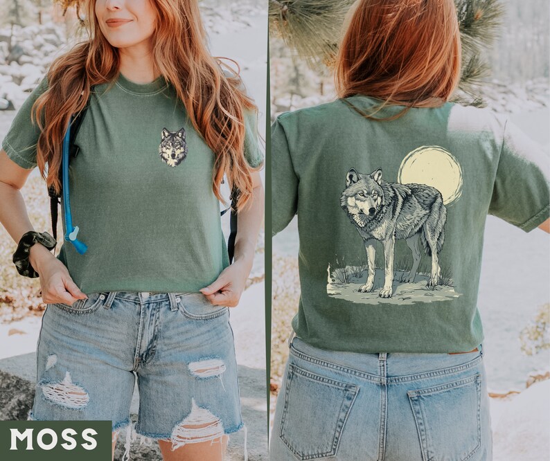May include: A green t-shirt with a wolf graphic on the front and a wolf graphic with a full moon on the back. The model is wearing the shirt with blue denim shorts.